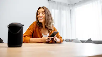 Lächelnde Frau am Tisch hält ein Smartphone und blickt zu einem schwarzen Smart Speaker.