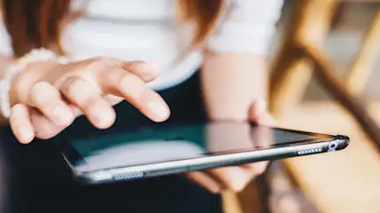 Eine Person tippt auf ein schwarzes Tablet. Im Hintergrund ist ein hölzerner Stuhl.