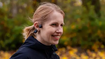 Eine lächelnde Frau trägt schwarze Kopfhörer und eine dunkle Jacke, im Hintergrund sind Bäume und gelbes Laub.