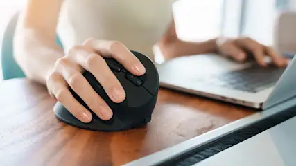 Eine Hand bedient eine schwarze, vertikale Computermaus auf einem Holztisch, neben einem teilweise sichtbaren Laptop.