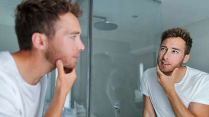 Ein Mann mit Drei-Tage-Bart blickt in einen Badezimmerspiegel.