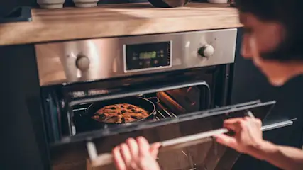 Eine Person öffnet einen silberfarbenen Ofen, in dem ein runder Kuchen auf einem Rost steht. Die Arbeitsplatte ist aus Holz.