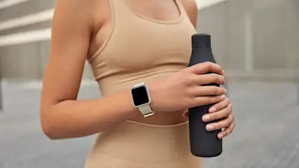 Eine Frau im Sport-BH hält eine schwarze Wasserflasche. Sie trägt eine Smartwatch mit einem hellen Armband.