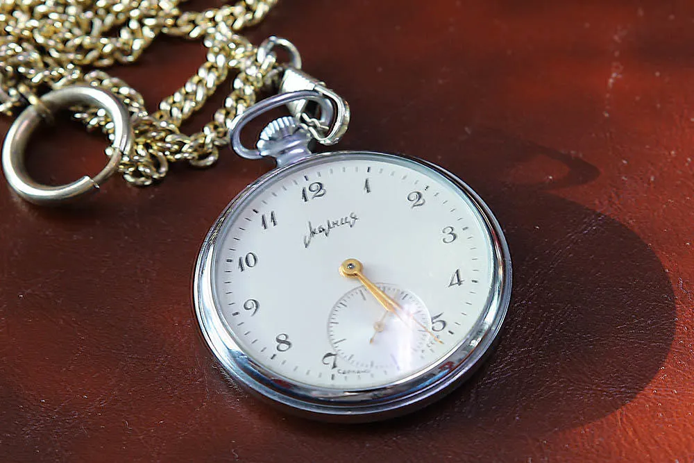 Eine silberne Taschenuhr mit goldener Kette liegt auf einer braunen Oberfläche. Die Uhr zeigt die Zeit an.