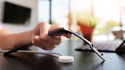 Eine Hand hält eine Smartwatch über einem weißen, runden Ladegerät auf einem dunklen Tisch, mit einem Laptop im Hintergrund.