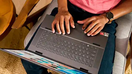 Eine Person sitzt und benutzt einen Laptop mit sichtbarer Tastatur und Bildschirm. Eine Uhr ist am Handgelenk der Person befestigt.