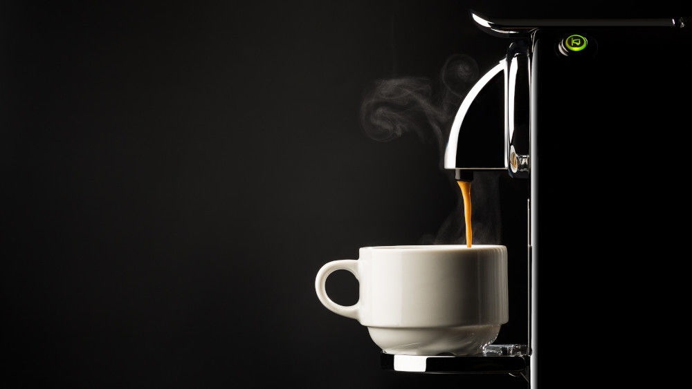 Eine Kaffeemaschine lässt dampfenden Kaffee in eine Tasse vor schwarzem Hintergrund