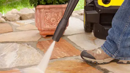 Eine Person reinigt mit einem Hochdruckreiniger eine Steinterrasse, neben einem Blumentopf und einem gelben Gerät.