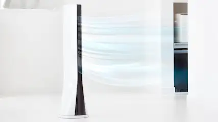 Ein Turmventilator in Weiß und Schwarz steht in einem hellen Raum und erzeugt einen sichtbaren Luftstrom.