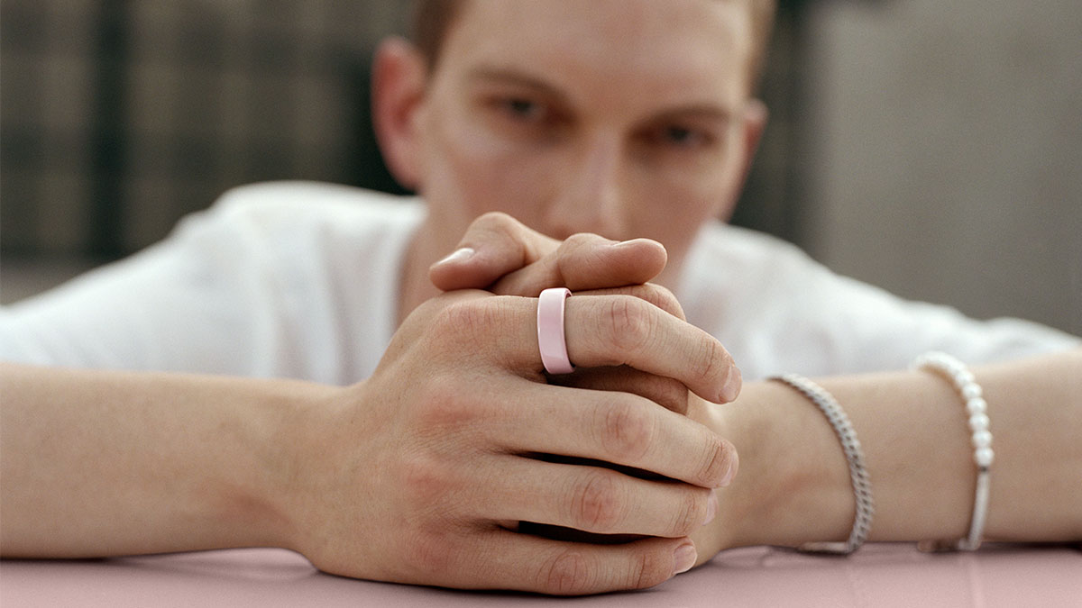 Person mit weißem T-Shirt lehnt gefaltete Hände auf rosa Fläche und trägt rosa Oura Ring 4 am Zeigefinger.