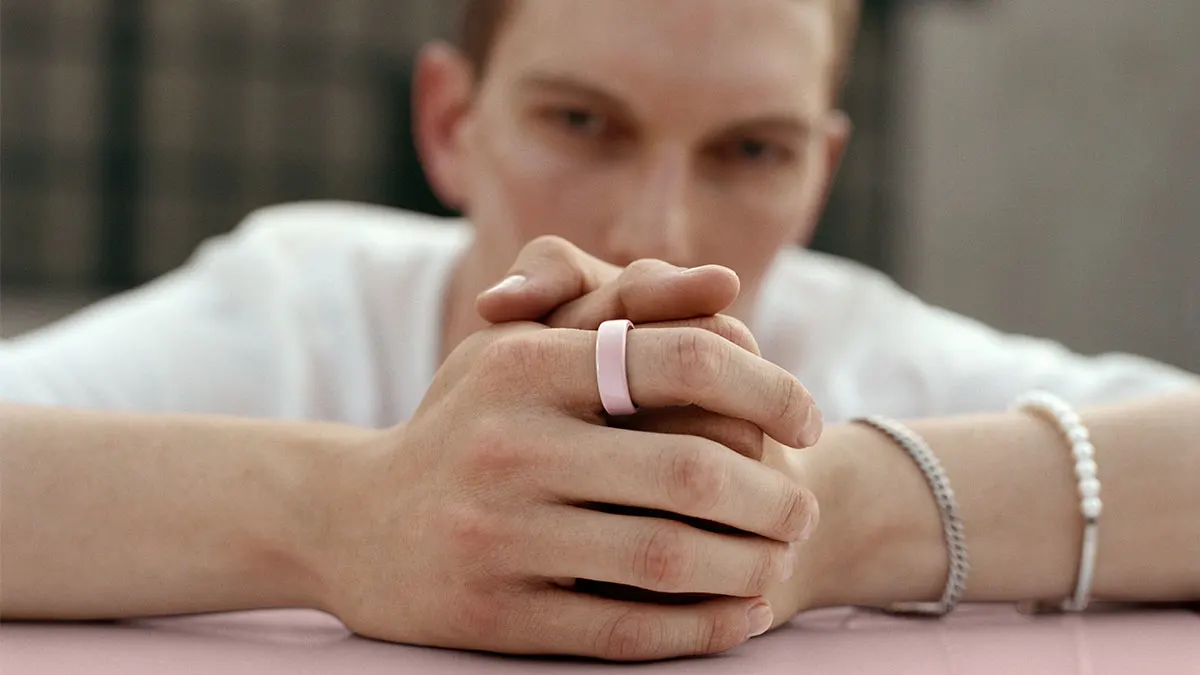 Person mit weißem T-Shirt lehnt gefaltete Hände auf rosa Fläche und trägt rosa Oura Ring 4 am Zeigefinger.