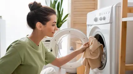 Frau in grünem T-Shirt entnimmt Wäsche aus einer weißen Waschmaschine in einem hellen Raum.