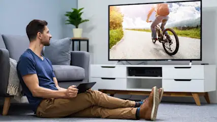 Mann sitzt vor TV mit Radfahrer-Szene, hält Tablet. Graues Sofa, weißes Lowboard, Zimmerpflanze im Hintergrund.