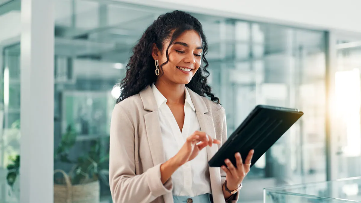Lächelnde Frau mit lockigen Haaren im beigen Blazer tippt auf einem schwarzen Tablet im hellen Büro.
