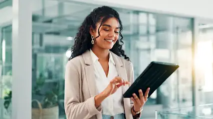 Lächelnde Frau mit lockigen Haaren im beigen Blazer tippt auf einem schwarzen Tablet im hellen Büro.
