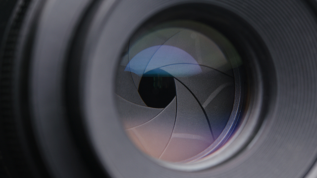 Nahaufnahme einer Kameraobjektivöffnung mit grauen Blendenlamellen um ein schwarzes Loch.
