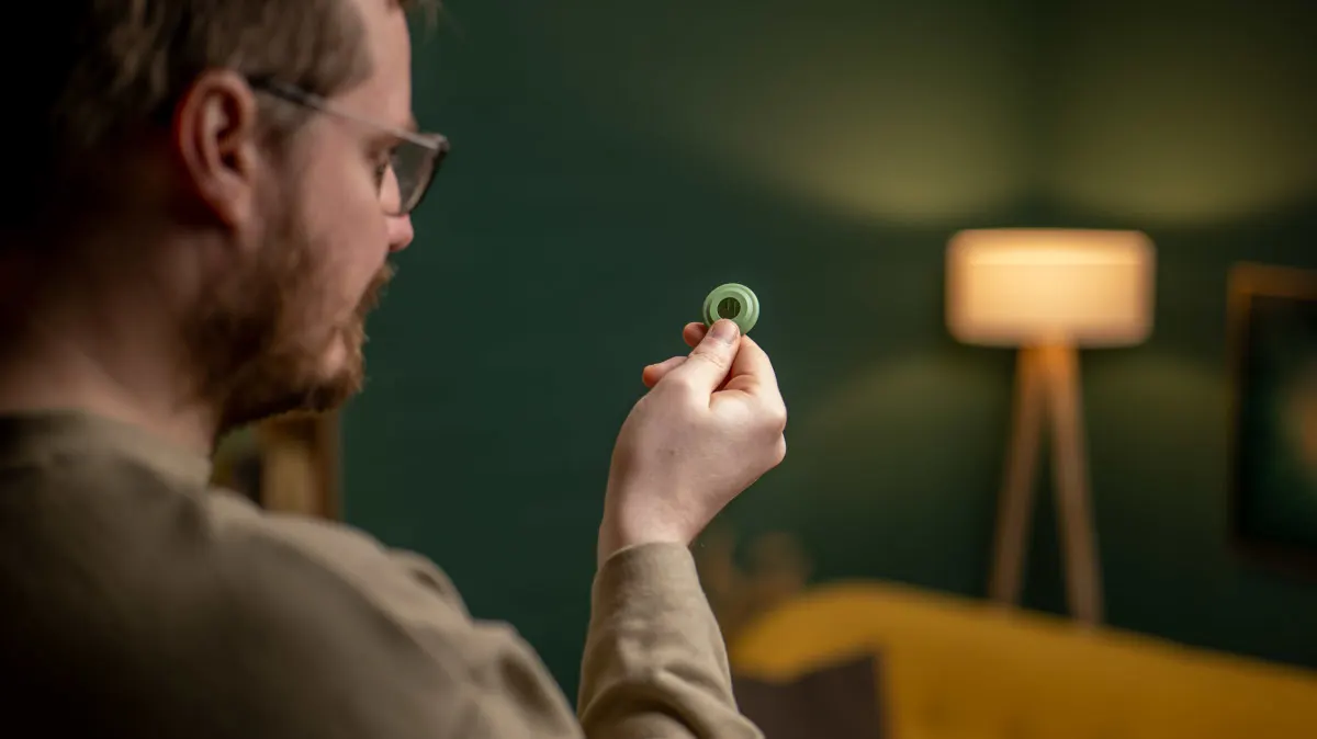 Ein Mann mit Brille hält einen kleinen, grünen Ring in der Hand. Im Hintergrund sind eine Lampe und eine grüne Wand zu sehen.