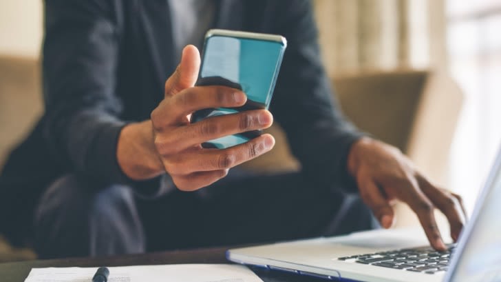 Ein Businessman sitzt auf dem Sofa. Sein Gesicht ist nicht zu sehen. Er hält ein Smartphone in der rechten Hand. Mit der linken Hand tippt er auf ein Laptop, das vor ihm auf einem niedrigen Couchtisch steht.