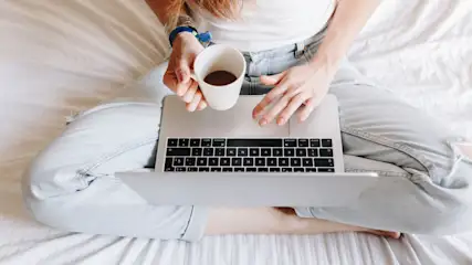 Eine Person sitzt im Schneidersitz auf einem Bett mit Laptop, hält eine Tasse und tippt.