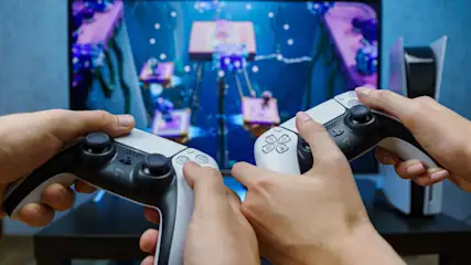 Zwei Hände halten Controller vor einem TV-Bildschirm, der ein Videospiel mit blauen und lila Elementen zeigt. Eine Spielkonsole steht rechts.