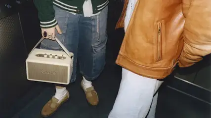 Zwei Personen mit Jeans, Lederjacke und Loafers im Aufzug. Eine Person trägt einen tragbaren Marshall-Lautsprecher.