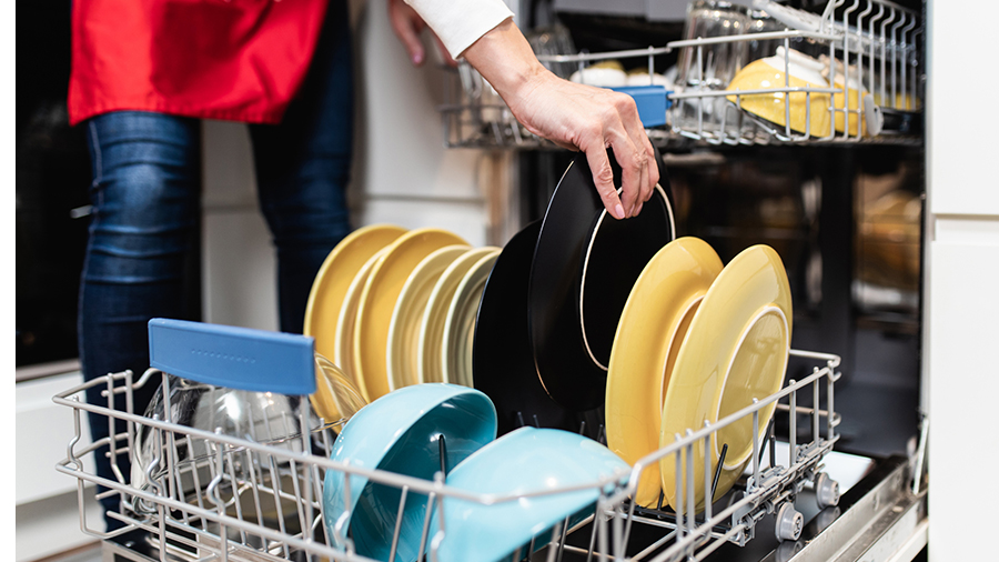Eine Person belädt einen Geschirrspüler mit gelben Tellern, schwarzen Tellern und blauen Schüsseln in einem Drahtgestell.