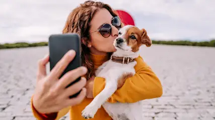 Eine Frau mit Sonnenbrille küsst einen kleinen Hund mit braunem Halsband, während sie ein schwarzes Handy hält. Im Hintergrund sind ein See und Bäume.