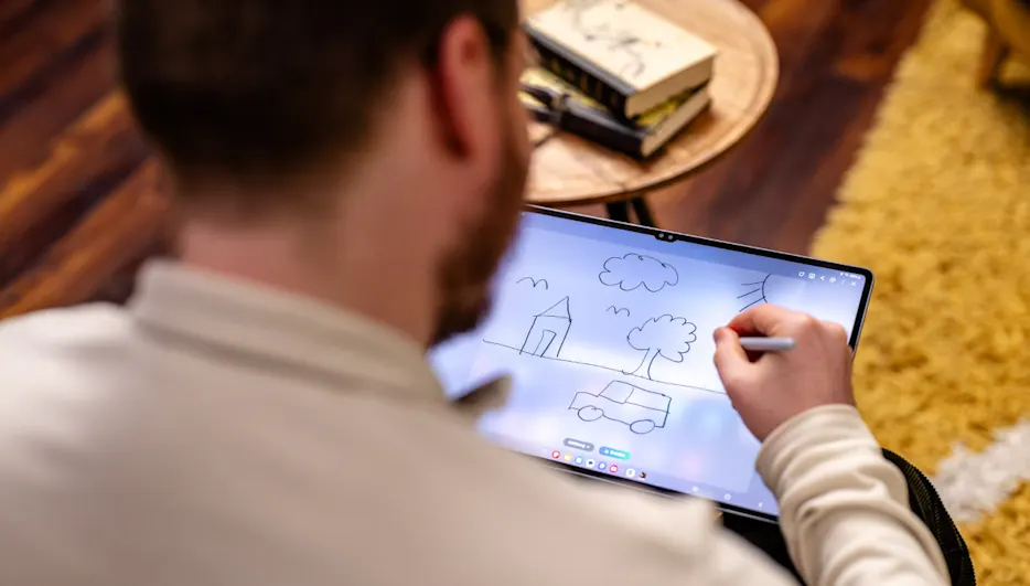 Eine Person zeichnet mit einem Stift auf einem Tablet-Bildschirm ein Haus, Wolken, einen Baum und ein Auto. Bücher auf einem Tisch.