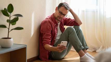Ein Mann sitzt auf dem Boden mit einem Handy in der Hand und fasst sich währenddessen an den Kopf.