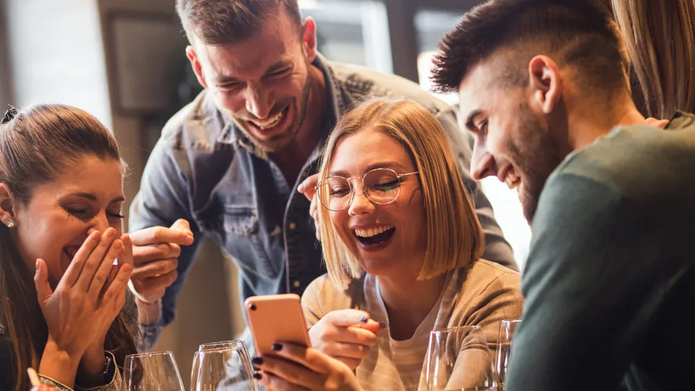 Vier lachende Personen um einen Tisch; eine Frau hält ein rosa Smartphone. Weingläser stehen auf dem Tisch.