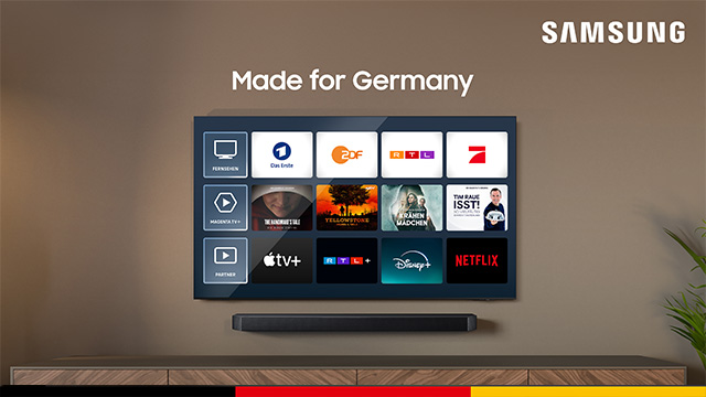 Ein Samsung-Fernseher hängt an einer Wand über einem Sideboard. Auf dem Bildschirm sind App-Logos zu sehen. Eine Pflanze steht rechts.