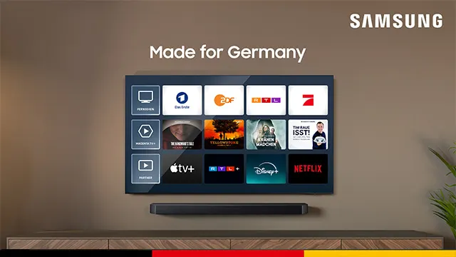 Ein Samsung-Fernseher hängt an einer Wand über einem Sideboard. Auf dem Bildschirm sind App-Logos zu sehen. Eine Pflanze steht rechts.