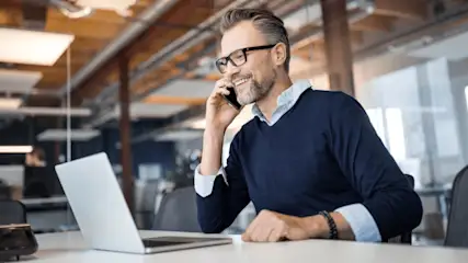 Ein Mann mit Brille telefoniert am Schreibtisch mit Laptop in einem Büro.