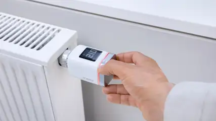 Eine Hand bedient ein weißes Bosch Heizkörperthermostat an einem weißen Heizkörper vor einer weißen Wand.