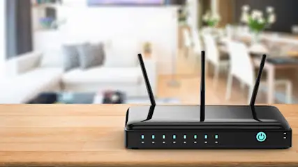 Ein Router mit drei Antennen steht auf einem Holztisch. Im Hintergrund sind eine Couch und ein Esstisch verschwommen zu sehen.