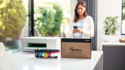 Auf einem Tisch stehen ein Drucker, Tintenpatronen und ein Paket von EcoPro. Im Hintergrund schaut eine Frau auf ihr Smartphone.