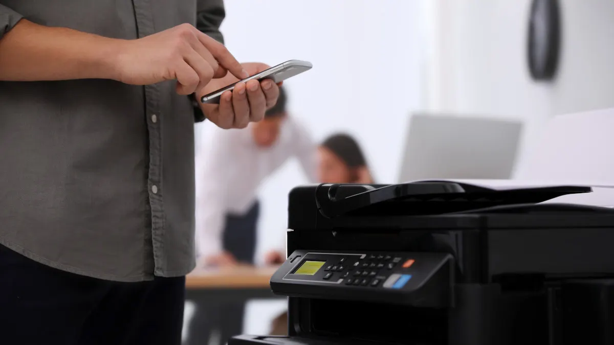In einem Büro steht eine Person mit ihrem Smartphone vor einem Drucker.