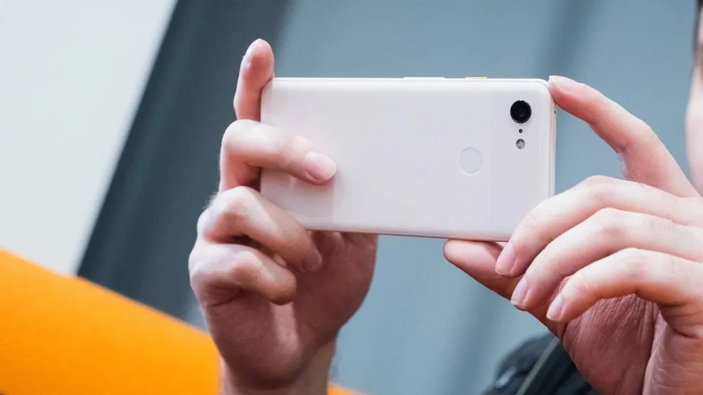 Eine Person hält ein weißes Smartphone mit einer schwarzen Kamera in horizontaler Ausrichtung mit beiden Händen fest.