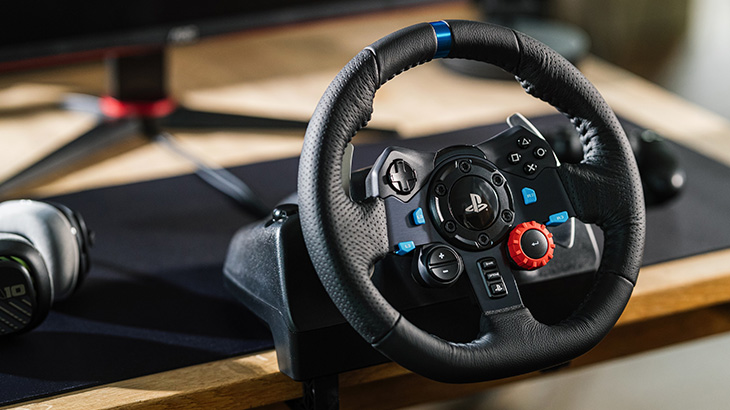 Ein schwarzes Lenkrad mit blauen und roten Knöpfen steht auf einem Schreibtisch mit einer schwarzen Matte, einem Monitor und Kopfhörern.