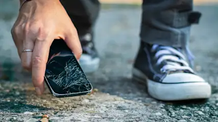 Eine Hand hält ein zerbrochenes schwarzes Handy über dem Boden. Im Hintergrund sind Schuhe und graue Jeans zu sehen.
