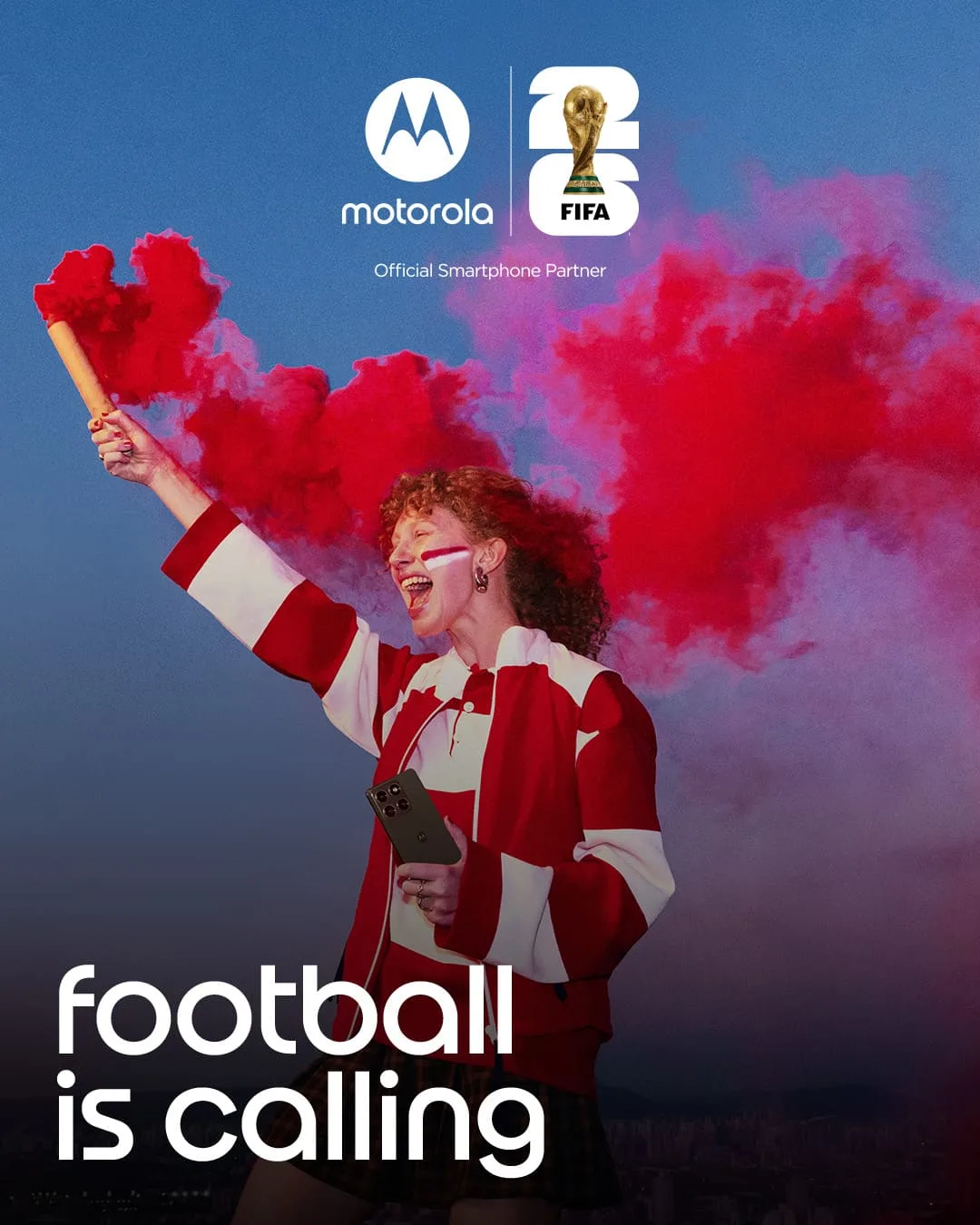 Fußballfan in rot-weißer Jacke jubelt mit rotem Rauchfackel in der Hand und hält ein Motorola‑Smartphone; darüber Motorola- und FIFA-Logos mit dem Hinweis „Official Smartphone Partner“, unten der Schriftzug „football is calling“.