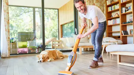 Ein Mann staubsaugt den Holzboden. Ein Hund liegt daneben. Im Hintergrund sind Fenster, ein Regal und Möbel zu sehen.