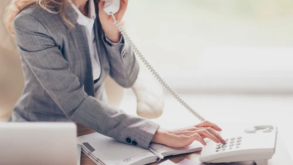 Eine Person in einem grauen Blazer telefoniert und tippt auf ein weißes Tischtelefon neben einem Notizbuch.