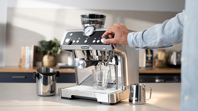 Ein Mann dreht an einem Knopf einer Delonghi Siebträgermaschine, während eine leeres Glas darunter steht.