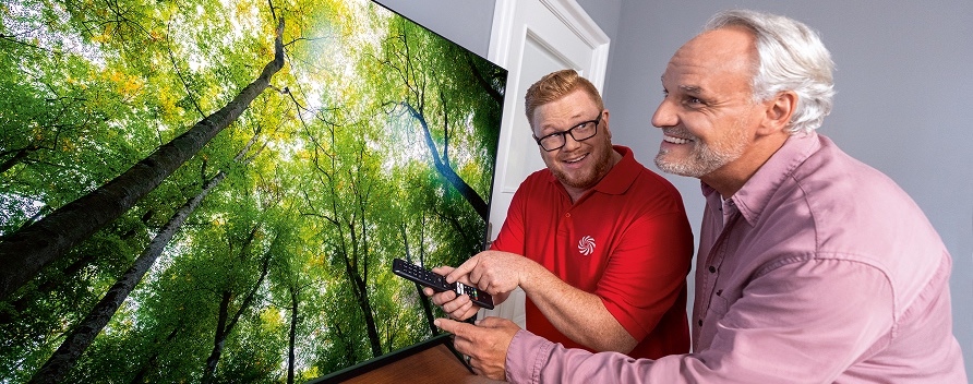 Techniker im roten Poloshirt erklärt einem älteren Mann die Fernbedienung eines Fernsehers.