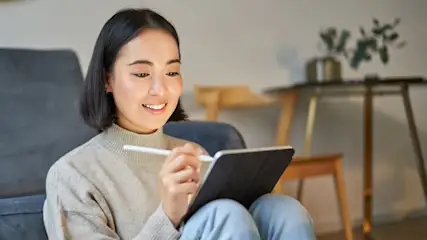 Frau sitzt in einem grauen Sessel, mit einem Stift auf einem Tablet. Im Hintergrund ein Tisch mit einer Pflanze.