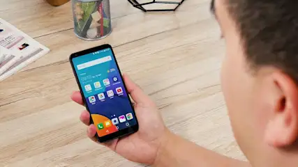 Eine Person hält ein Smartphone mit blauem Bildschirm über einem Holztisch. Bürobedarf im Hintergrund.