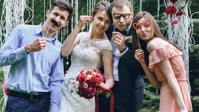 Eine Hochzeitsgesellschaft posiert mit Requisiten wie einem falschen Schnurrbart und Brillen vor einem dekorativen Hintergrund.