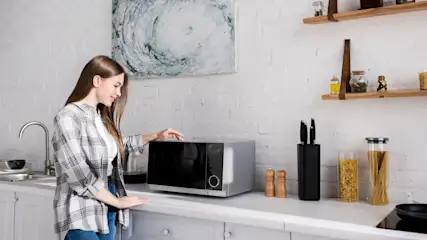 Frau steht in einer Küche und berührt eine Mikrowelle. Regale halten Gläser und Gewürze. Ein abstraktes Gemälde hängt an der Wand.