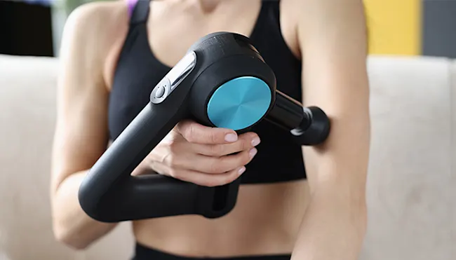 Frau in Sportbekleidung massiert ihren Arm mit einem schwarzen Massagegerät mit blauem Akzent.